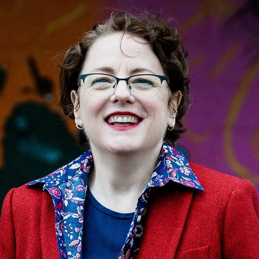 Paula McConnell, the founder of Seva Digital, looks forward smiling while wearing a red jacket and floral blouse.